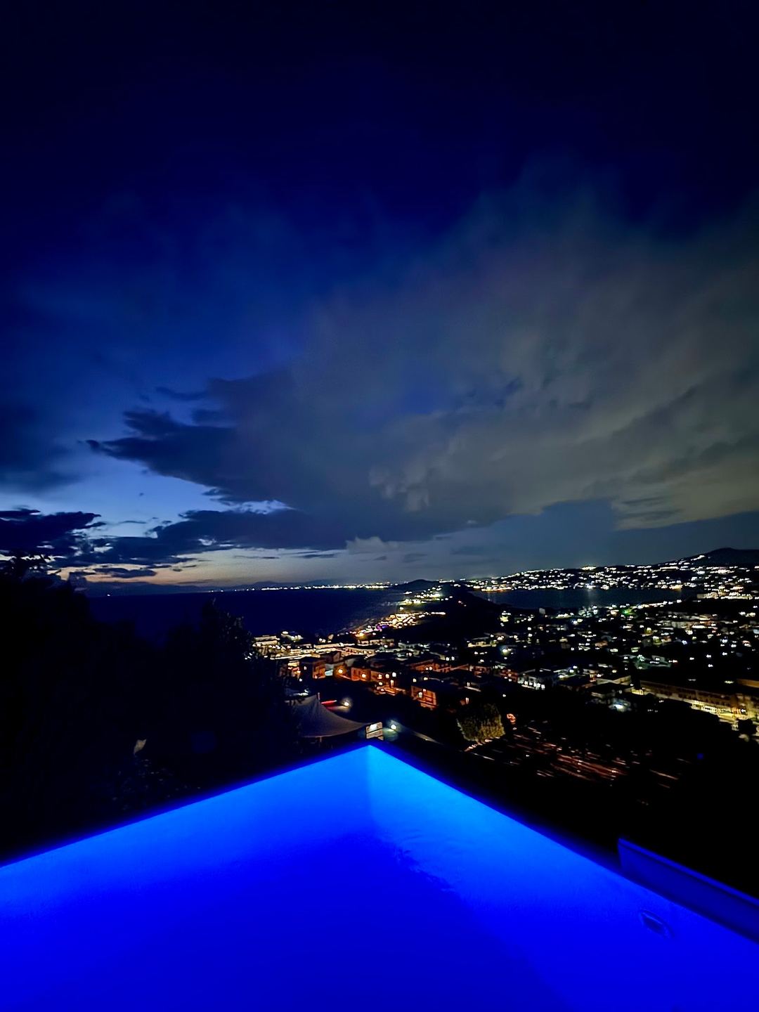 An illuminated infinity pool overlooks a coastal town at twilight with mountains and city lights in the background.