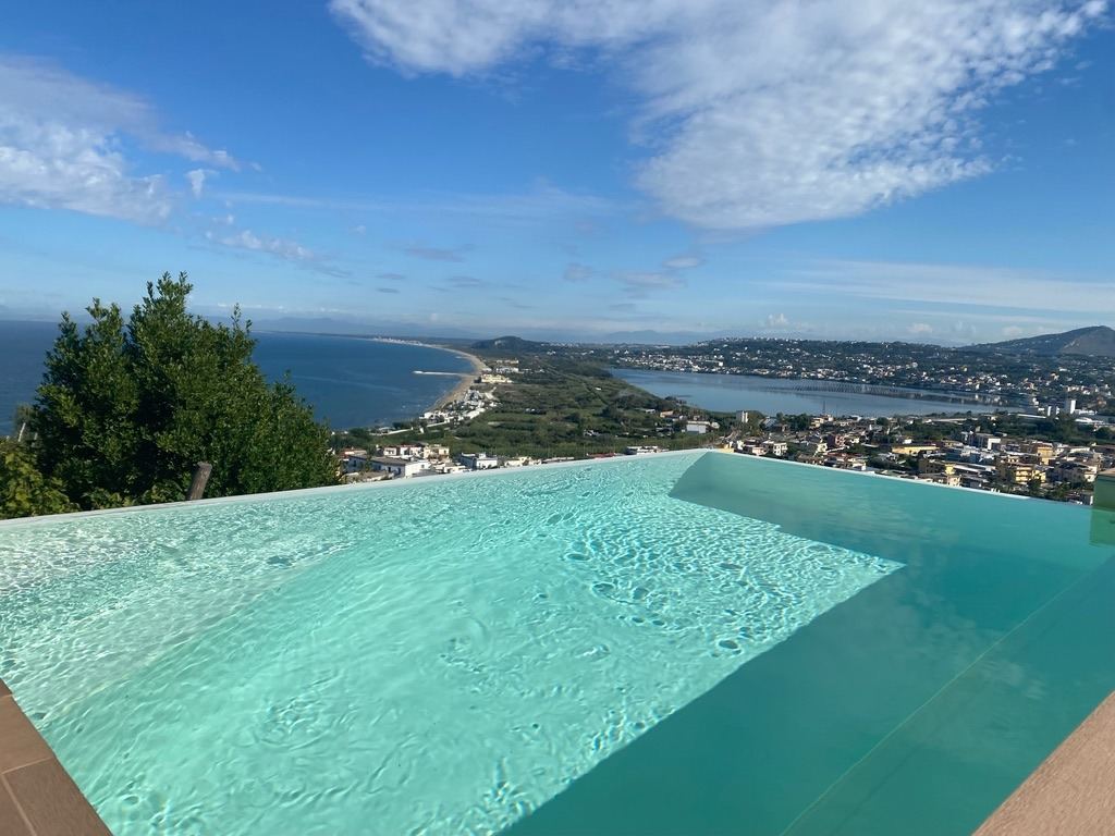 An infinity pool with crystal-clear turquoise water overlooks a Mediterranean coastal town and bay below.