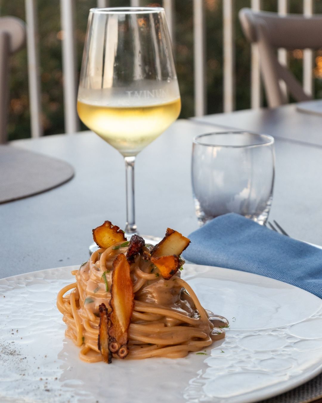 An elegantly plated pasta dish with roasted vegetables topped with a caramelized garnish, paired with a glass of white wine on an outdoor dining table.