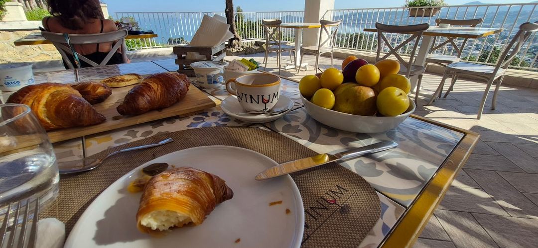Breakfast spread on a seafront terrace featuring fresh croissants, fruits, and coffee with panoramic coastal views.